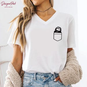 Puede incluir: Camiseta blanca con cuello en V con un dise&ntilde;o negro de un perezoso asomando por un bolsillo. El bolsillo est&aacute; delineado en negro. La camiseta tiene mangas cortas y es de material suave. El texto "Designer Park Special Designs" est&aacute; en la esquina superior izquierda.
