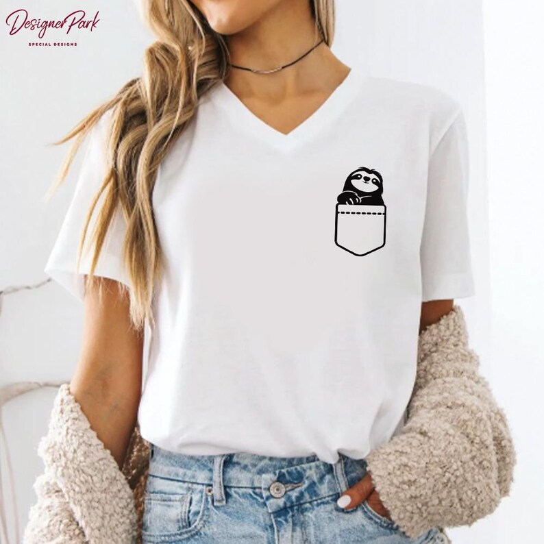 Puede incluir: Camiseta blanca con cuello en V con un dise&ntilde;o negro de un perezoso asomando por un bolsillo. El bolsillo est&aacute; delineado en negro. La camiseta tiene mangas cortas y es de material suave. El texto "Designer Park Special Designs" est&aacute; en la esquina superior izquierda.
