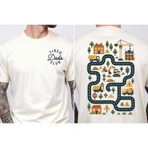 May include: Cream-colored t-shirt with the text "Tired Dads Club" on the front. The back features a colorful construction site design with trucks, houses, trees, and a crane. The design includes a winding road and construction vehicles.