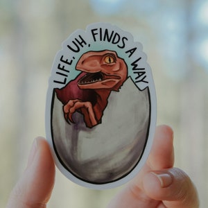 May include: A sticker featuring a cartoon raptor hatching from an egg. The raptor is red and brown with an orange eye. The sticker has the text "LIFE, UH, FINDS A WAY" in black lettering.
