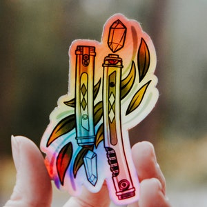 May include: A holographic sticker featuring two colorful wands with crystals at the top, surrounded by stylized leaves. The sticker has a rainbow gradient background.