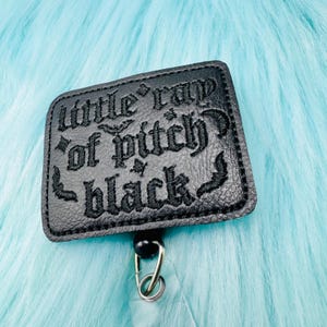 May include: Black badge reel with the text "little rap of pitch black" in gothic lettering. The badge is rectangular with rounded corners and a textured surface. A black bead and metal clip are attached.