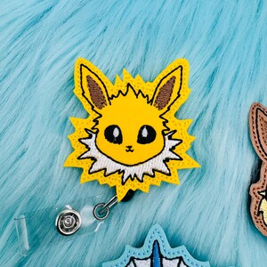 K&ouml;nnte beinhalten: Ein gelbes und wei&szlig;es gesticktes Abzeichen mit einem Pok&eacute;mon-Charakter, Eevee, mit einem durchsichtigen, einziehbaren Ausweishalter aus Kunststoff.