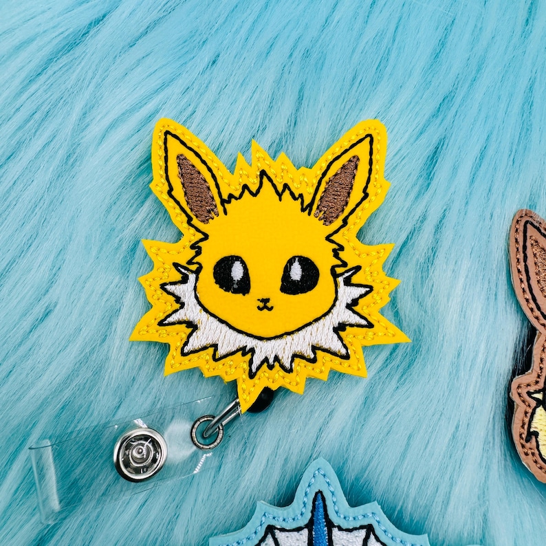 K&ouml;nnte beinhalten: Ein gelbes und wei&szlig;es gesticktes Abzeichen mit einem Pok&eacute;mon-Charakter, Eevee, mit einem durchsichtigen, einziehbaren Ausweishalter aus Kunststoff.