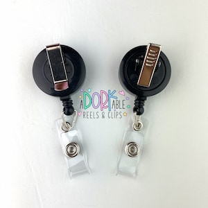 May include: Two black retractable badge reels with silver clips and clear plastic badge holders. The reels have the text "A DORK ABLE REELS & CLIPS" in a colorful, whimsical font.