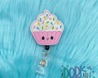May include: A pink cupcake-shaped badge reel with a smiling face, white frosting, and colorful sprinkles. The badge reel has a retractable cord and a clear plastic clip. The text "A DORK ABLE REELS & CLIPS" is in the bottom right corner.