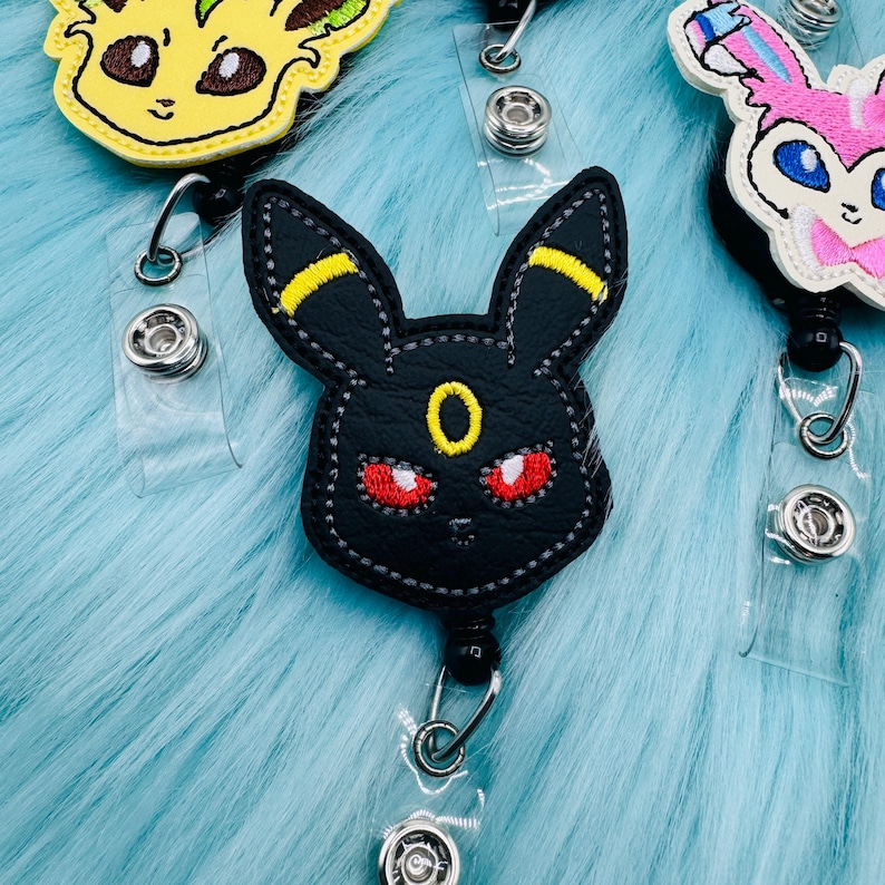 K&ouml;nnte beinhalten: Ein schwarzer Leder-Badgehalter mit einem gestickten Pokemon-Charakter in Schwarz und Gelb. Der Charakter hat rote Augen und einen gelben Ring um den Hals.