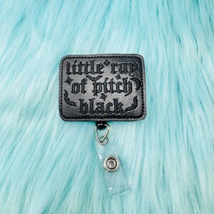 May include: Black rectangular badge reel with the text "little ray of pitch black" in gothic lettering. The badge reel has a clear plastic strap and a metal clip. The background is a light blue, fluffy material.