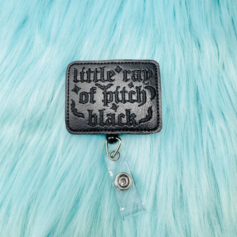 May include: Black rectangular badge reel with the text "little ray of pitch black" in gothic lettering. The badge reel has a clear plastic strap and a metal clip. The background is a light blue, fluffy material.