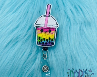 May include: An embroidered badge reel featuring a bubble tea cup with a rainbow-colored drink, black tapioca pearls, a pink straw, and a smiling face. The design is outlined in white and attached to a black retractable clip mechanism.