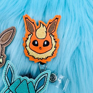 K&ouml;nnte beinhalten: Orangefarbener und gelber gestickter Abzeichen eines Pokemon-Charakters mit einer einziehbaren Abzeichenrolle.