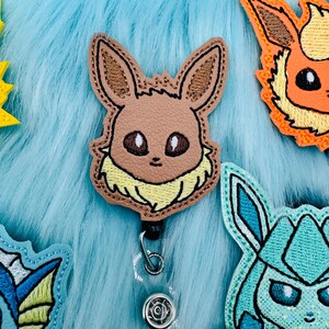 K&ouml;nnte beinhalten: Ein gesticktes Abzeichen mit dem Charakter Eevee in Braun und Gelb mit einem durchsichtigen, einziehbaren Ausweishalter aus Kunststoff.