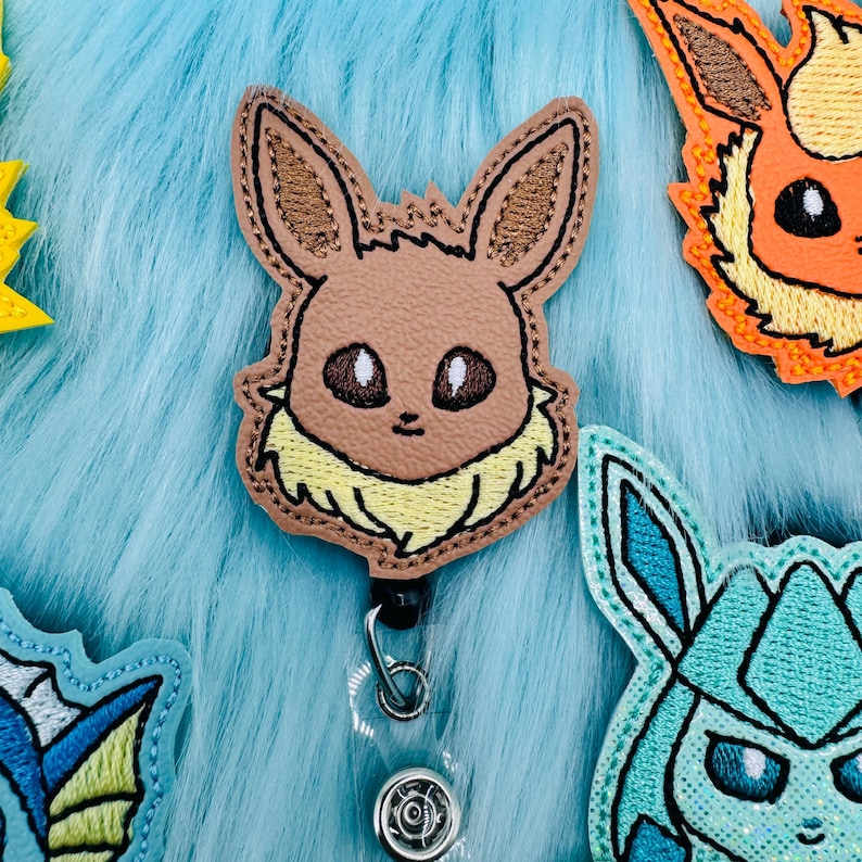 K&ouml;nnte beinhalten: Ein gesticktes Abzeichen mit dem Charakter Eevee in Braun und Gelb mit einem durchsichtigen, einziehbaren Ausweishalter aus Kunststoff.