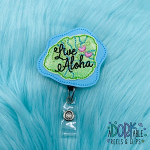 May include: A blue and green embroidered badge with the text "Live Aloha" and two pink hearts. The badge is attached to a clear plastic retractable badge holder.