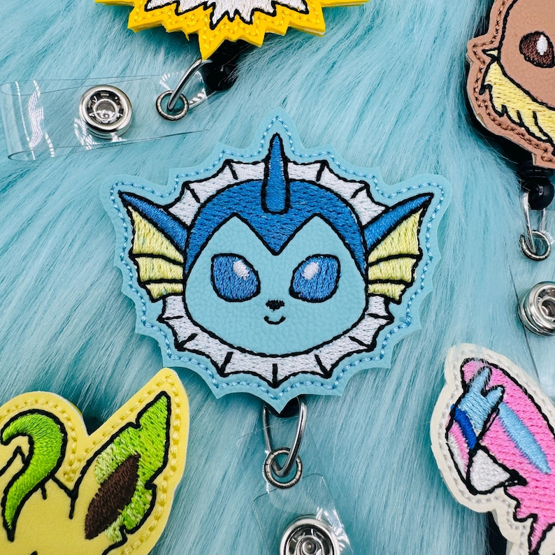 K&ouml;nnte beinhalten: Ein blaues und wei&szlig;es gesticktes Abzeichen mit einem Cartoon-Wasser-Pok&eacute;mon mit einem hellblauen K&ouml;rper, einem wei&szlig;en R&uuml;schen und gelben Flossen. Das Abzeichen ist an einem durchsichtigen Plastik-Badgehalter mit einem Metallclip befestigt.
