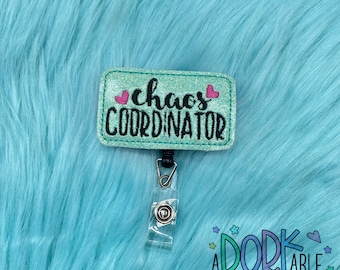 May include: A teal glitter badge reel with the words "chaos COORDINATOR" in black script and block letters. Two pink hearts are on the right side of the word "chaos". The badge reel has a clear plastic clip and a black retractable cord. The background is a fluffy, light blue material.