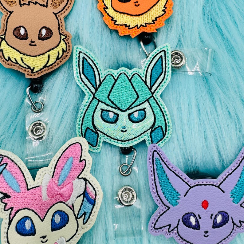 K&ouml;nnte beinhalten: F&uuml;nf gestickte Pok&eacute;mon-Charakter-Badge-Rollen. Die Rollen sind t&uuml;rkis, lila, rosa, braun und orange. Die Charaktere sind Eevee, Espeon, Umbreon, Vaporeon und Flareon.