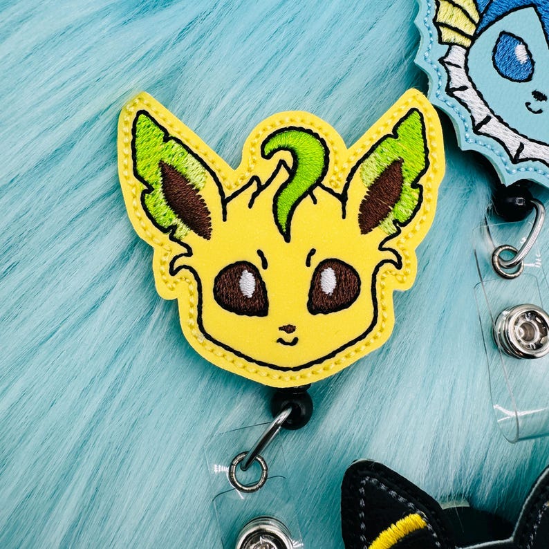 K&ouml;nnte beinhalten: Ein gelber gestickter Abzeichen mit einem gr&uuml;nen und braunen Blattdesign, das eine Comicfigur mit gro&szlig;en Augen und einem kleinen Mund zeigt. Das Abzeichen ist an einem einziehbaren Ausweishalter befestigt.
