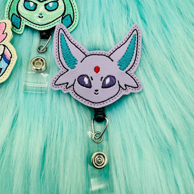 K&ouml;nnte beinhalten: Lila und t&uuml;rkisfarbener bestickter Badge-Rollenhalter mit einer Cartoon-Darstellung eines lilafarbenen Pok&eacute;mon mit einem roten Punkt auf der Stirn.