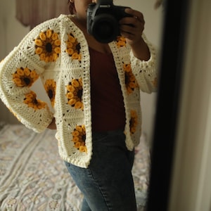 Pode incluir: Um casaco de crochê branco com motivos de girassóis amarelos. O casaco tem um decote em V e mangas compridas.