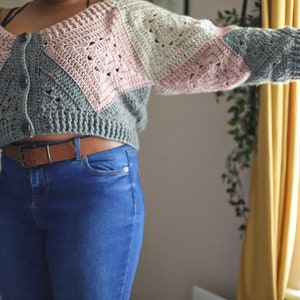 Puede incluir: Un cárdigan corto con cierre de botones, con un diseño de patchwork de cuadrados de crochet rosa, verde y blanco. El cárdigan se lleva sobre un vaquero azul.