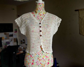 May include: White crochet top with a button-up front and short sleeves. The top is on a floral-patterned mannequin.