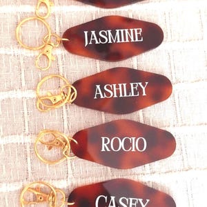 May include: Five brown tortoise shell keychains with gold key rings and clasps. Each keychain has a white oval tag with a name on it: Kayla, Jasmine, Ashley, Rocío, and Casey.