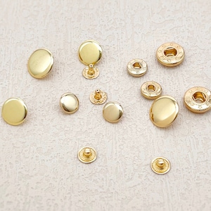 Puede incluir: Surtido de botones a presión de metal dorado en varios tamaños y estilos. Los botones a presión están dispuestos sobre una superficie beige claro.