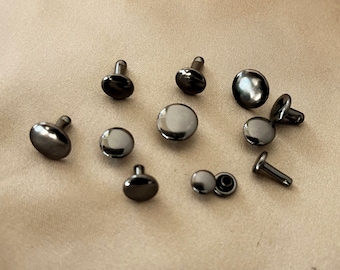 Può includere: Una collezione di dieci rivetti in metallo nero, alcuni con un perno e altri con un retro piatto. I rivetti sono sparsi su una superficie di tessuto marrone chiaro.