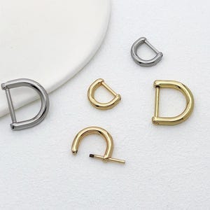 May include: A selection of metal D-ring buckles in silver and gold hues. The buckles are of varying sizes and shapes, with some having a rounded D form and others a more squared design. The buckles are displayed on a white surface.