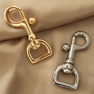 May include: Two metal swivel snap hooks, one gold and one silver, are shown against a neutral backdrop. The hooks feature a spring-loaded clasp and a D-ring for attachment, suitable for crafting and hardware projects. The background is a light beige.