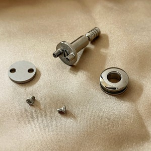 May include: A set of silver hardware for a project. The set includes a small round plate with two holes, a round ring with a hole in the centre, a small screw, a larger screw, and a silver piece with a small knob and two holes.