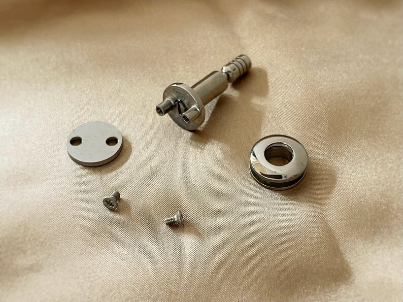 May include: A set of silver hardware for a project. The set includes a small round plate with two holes, a round ring with a hole in the centre, a small screw, a larger screw, and a silver piece with a small knob and two holes.