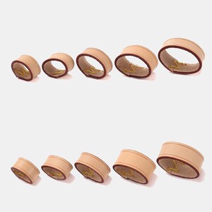 Peut inclure: Dix anneaux en cuir circulaires dispos&eacute;s en deux rang&eacute;es de cinq, variant de taille du plus petit au plus grand. Chaque anneau pr&eacute;sente un ext&eacute;rieur beige clair, une doublure int&eacute;rieure marron fonc&eacute; et une couture en zigzag jaune visible.