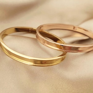 May include: Two gold-coloured bangles, one slightly overlapping the other, rest on a satin-like, beige surface. The bangles have a smooth, polished finish, reflecting light. The overall aesthetic is elegant and minimalist.