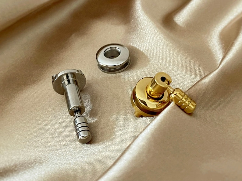May include: Two metal hardware pieces, one silver and one gold. The silver piece is a small, round, flat washer with a hole in the centre. The gold piece is a small, round, flat washer with a hole in the centre and a small, gold, metal knob attached to the side.