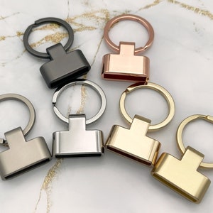 Op de afbeelding: Zes sleutelhangers met verschillende gekleurde metalen afwerkingen. De sleutelhangers hebben een ronde ring en een rechthoekig metalen label. De kleuren zijn zilver, goud, rosé goud, zwart en twee tinten grijs.