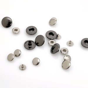 May include: A collection of black and silver metal snap fasteners. The fasteners are scattered on a white surface.
