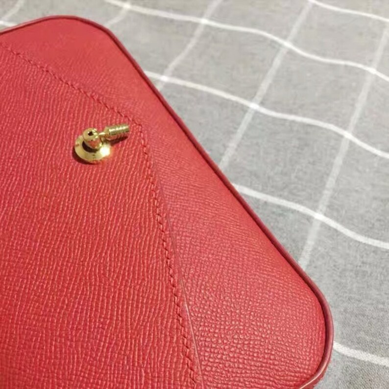 May include: Close-up of a red leather handbag with a gold metal clasp. The bag has a textured surface and a stitched seam.