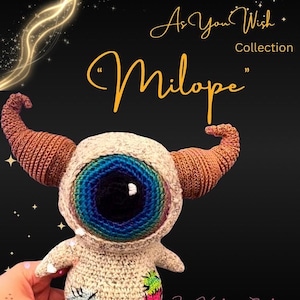 May include: A crocheted doll with a large, multi-coloured eye and brown horns. The doll is beige with patches of colour on its body. The text "As You Wish Collection" and "Milope" are displayed on a black background.