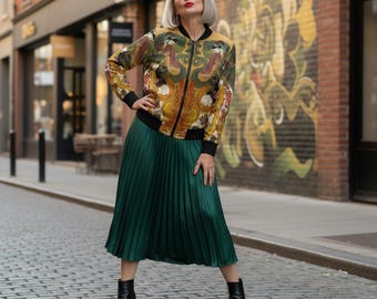 Puede incluir: Una mujer con una chaqueta bomber colorida, una falda plisada verde y botines negros. La chaqueta presenta un diseño ornamentado con tonos dorados, marrones y verdes. La falda llega a la pantorrilla, y los botines tienen tacón cuadrado. El fondo es una calle.