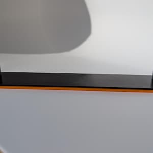 May include: A black rectangular shelf with orange trim and two black end pieces. The shelf is made of a glossy material and is designed to hold items. The shelf is on a white surface.