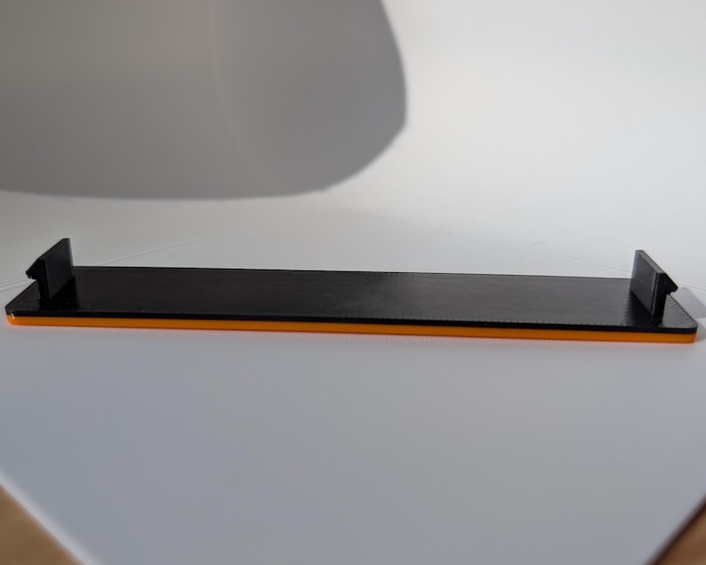 May include: A black rectangular shelf with orange trim and two black end pieces. The shelf is made of a glossy material and is designed to hold items. The shelf is on a white surface.