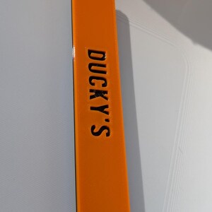 May include: An orange rectangular object with the text "DUCKY'S" in black. The object is a solid color and appears to be made of plastic or a similar material. The text is centered and vertically aligned.