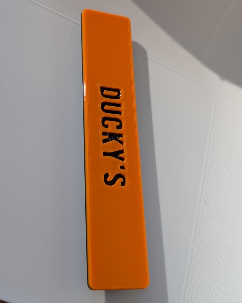 May include: An orange rectangular object with the text "DUCKY'S" in black. The object is a solid color and appears to be made of plastic or a similar material. The text is centered and vertically aligned.