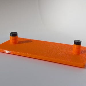 May include: An orange rectangular platform with a textured surface, featuring two small cylindrical posts. The posts are black on top and orange below. The platform is approximately 8 inches long and 2 inches wide.