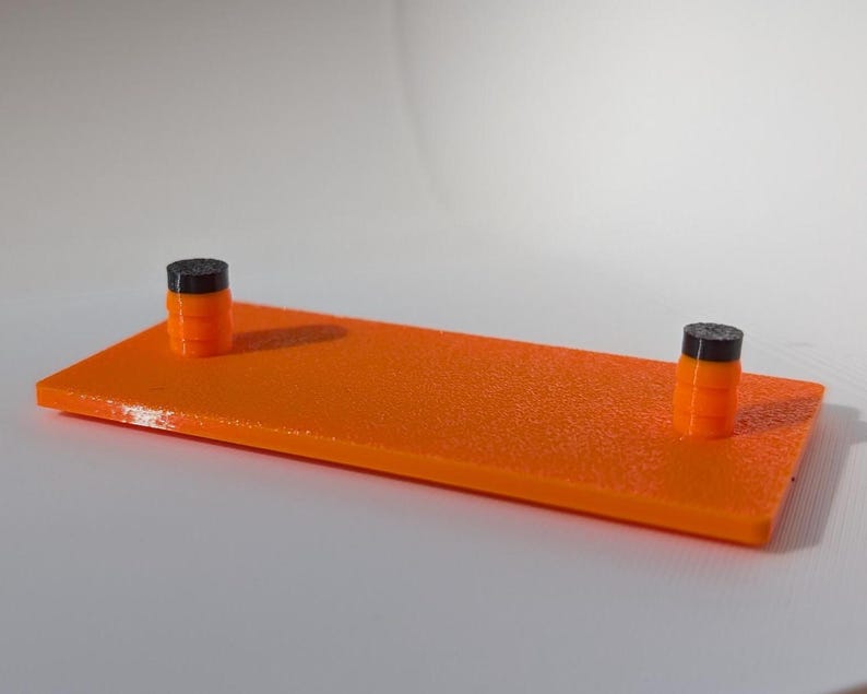 May include: An orange rectangular platform with a textured surface, featuring two small cylindrical posts. The posts are black on top and orange below. The platform is approximately 8 inches long and 2 inches wide.
