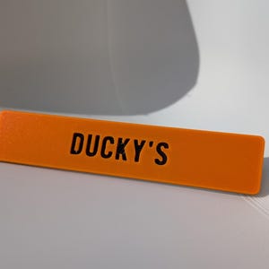 May include: An orange rectangular sign with the text "DUCKY'S" in black. The sign is made of a solid material and is resting on a white surface. The sign is a bright, eye-catching color.