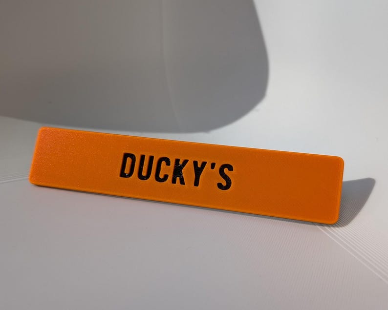 May include: An orange rectangular sign with the text "DUCKY'S" in black. The sign is made of a solid material and is resting on a white surface. The sign is a bright, eye-catching color.