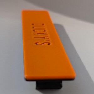 May include: An orange rectangular object with the word "DUCKY'S" embossed on its surface. The object has a black base and is set against a neutral background. The text is in a sans-serif font.